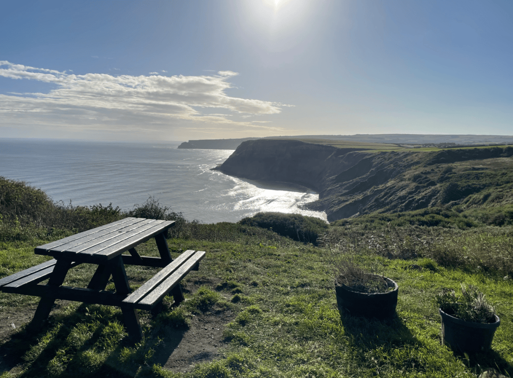 A picnic table overlooking a rugged coastal cliff and the ocean, with sun shining brightly in the sky.