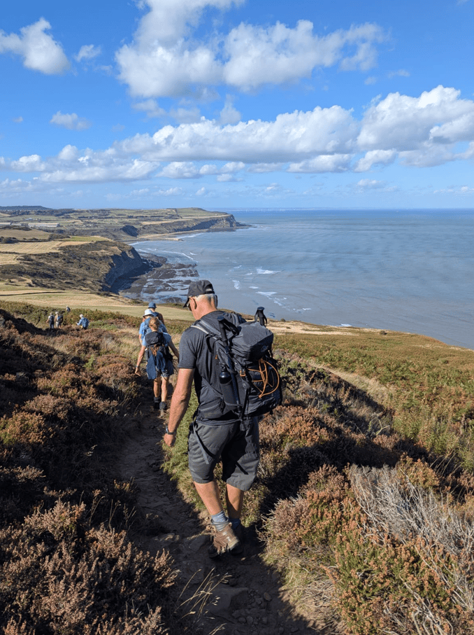 A group of hikers walking along a coastal path with cliffs and the sea in the background, under a blue sky with clouds.