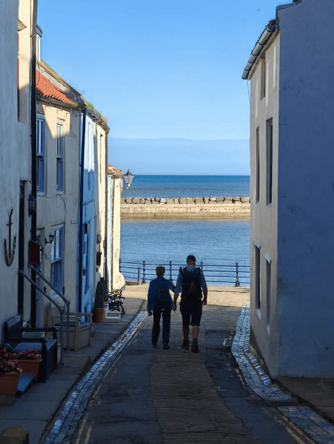 A couple holding hands walking down a narrow street between white and blue buildings, with a view of the sea in the background.