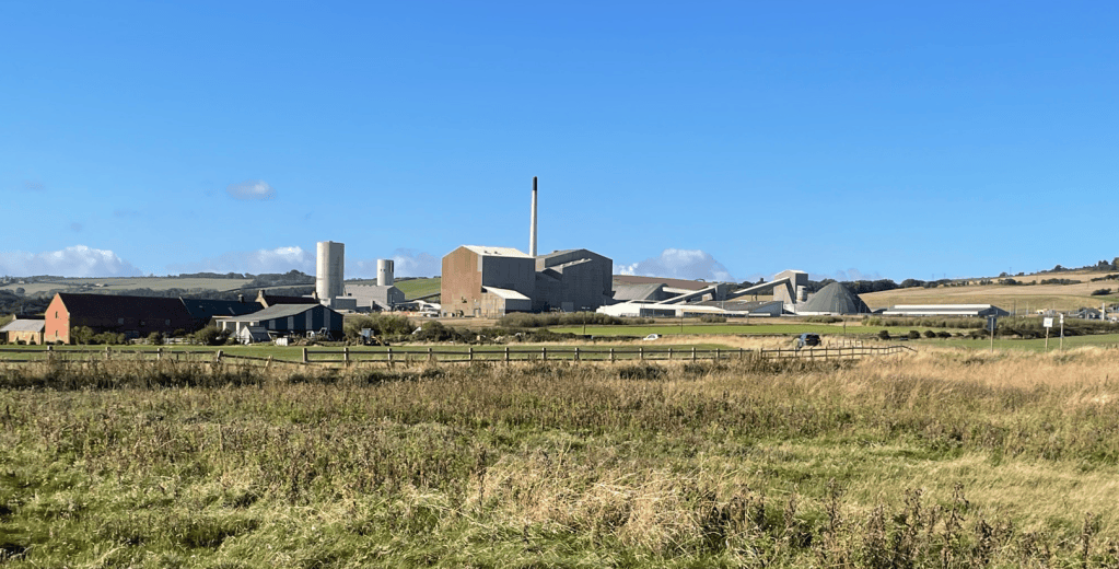 View of an industrial complex with various buildings and silos set against a blue sky, surrounded by fields and a few farmhouses.