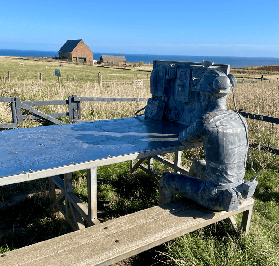 A weathered statue of a miner seated at a metal table, equipped with old-fashioned communication devices, set against a backdrop of green fields and a distant barn under a clear blue sky.