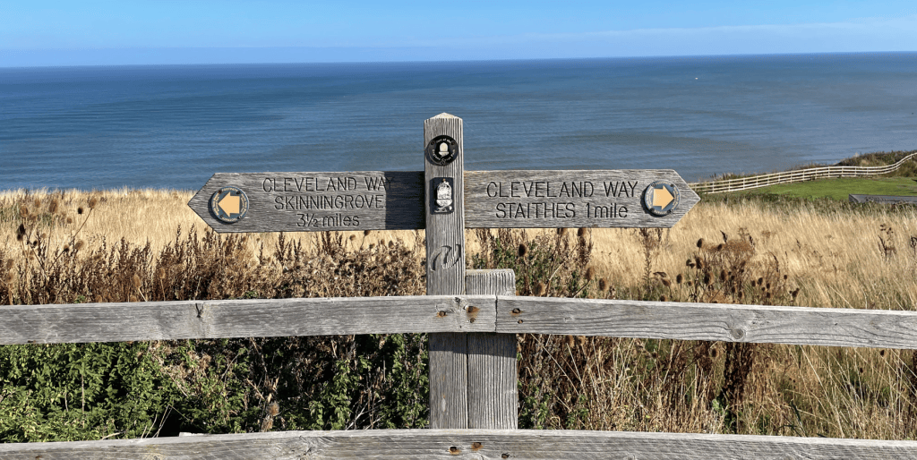 A wooden signpost displaying directions to Cleveland Way Skelton Green (3.5 miles) and Cleveland Way Staithes (1 mile), with a scenic view of the sea and grassland in the background.