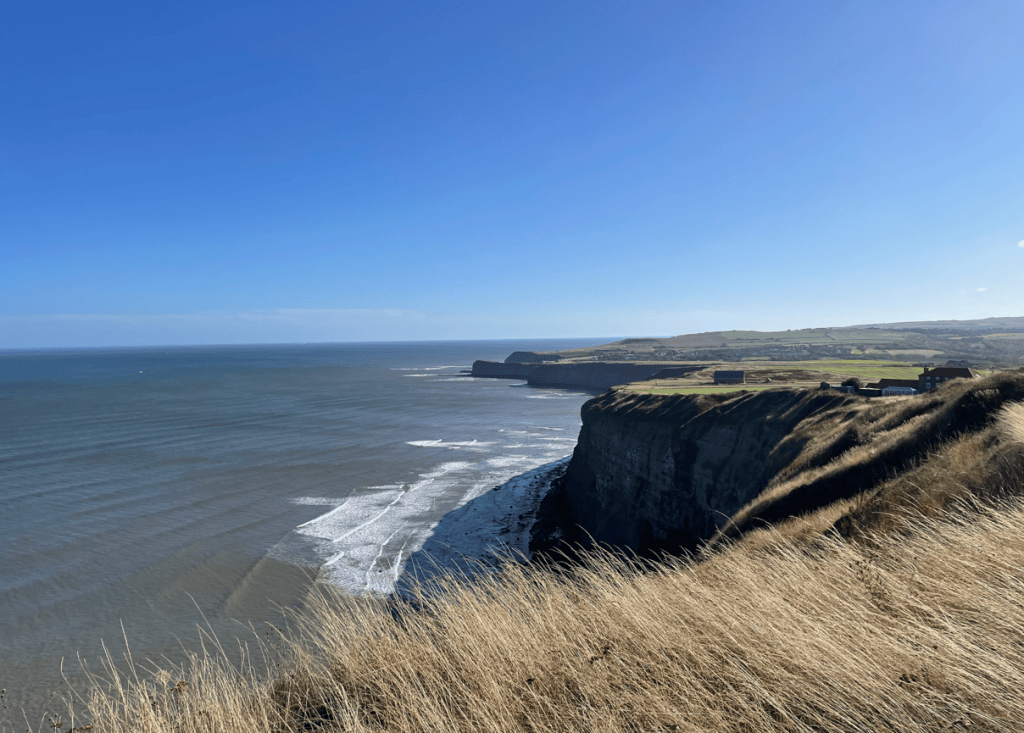 A scenic view of a rugged coastline with steep cliffs overlooking the ocean, under a clear blue sky. Gentle waves are lapping at the shore, while grassy vegetation adorns the cliff top.