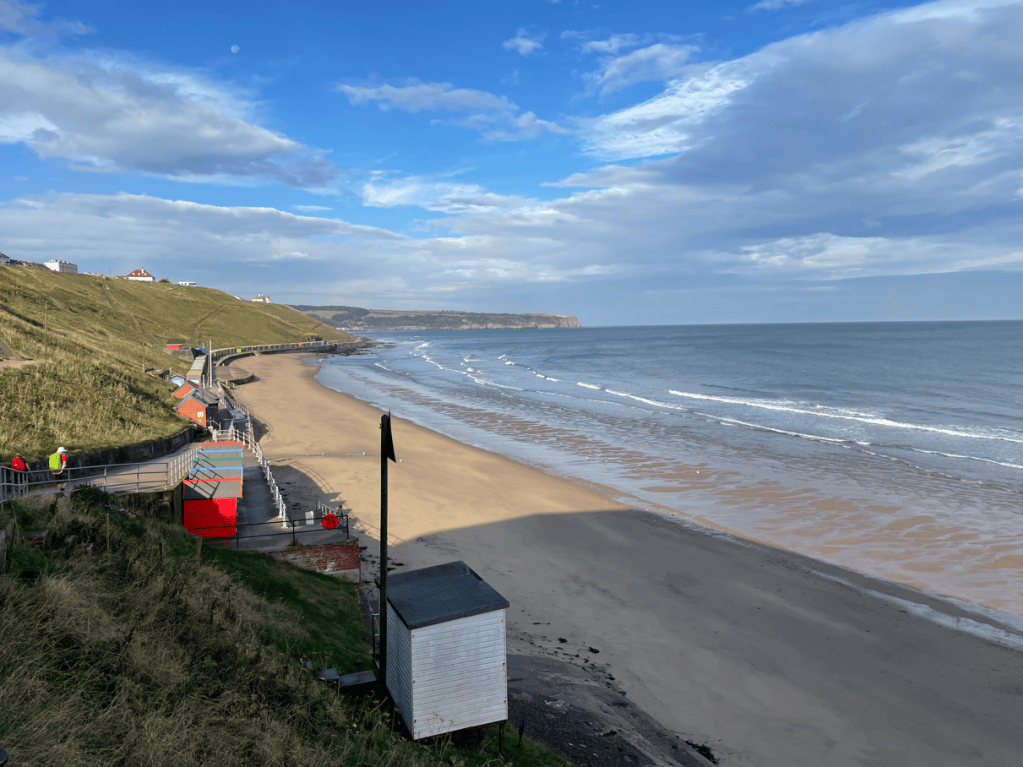 A scenic view of a beach with gentle waves, sandy shore, and a coastal path lined with colourful beach huts. The sky is partly cloudy with patches of blue.