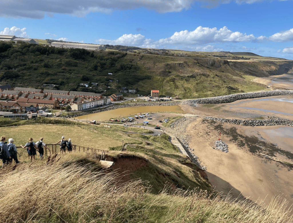 A scenic coastal view showing a village with houses, a river estuary, and a sandy beach. People are walking down a slope amidst tall grass, with cliffs and a clear blue sky in the background.