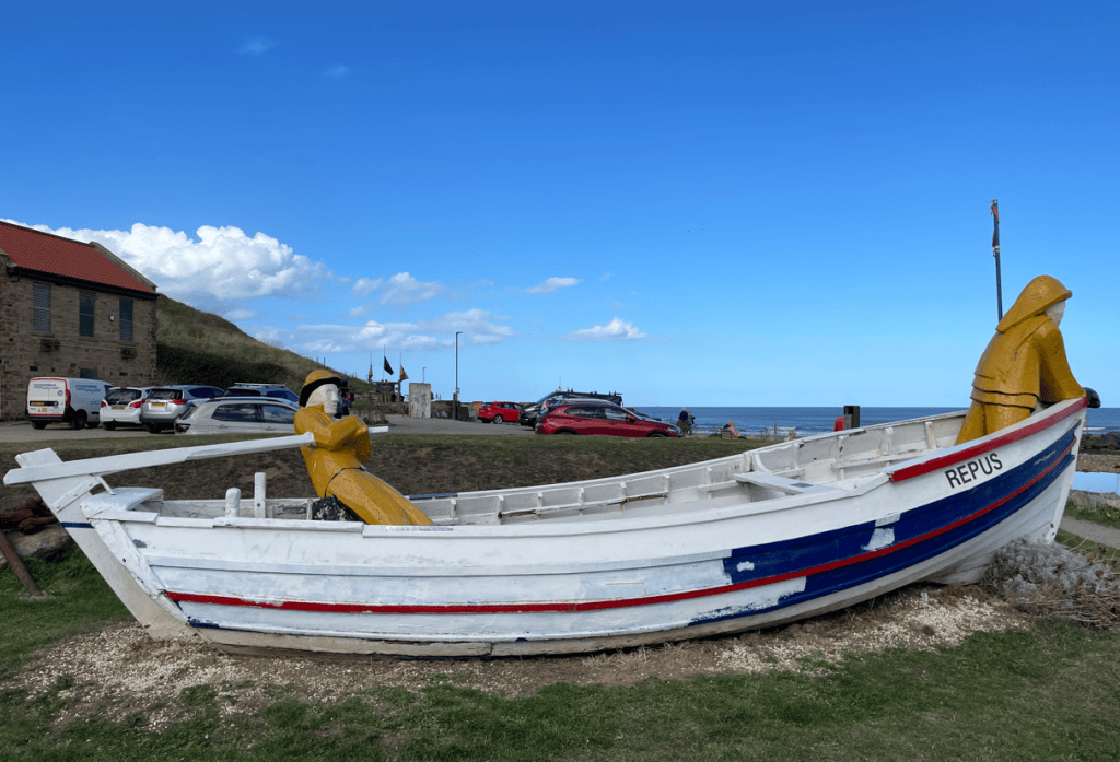 A decorative fishing boat named 'REPUS' with two colourful wooden sculptures of fishermen in bright yellow and traditional attire, situated on grassy land by the coast.