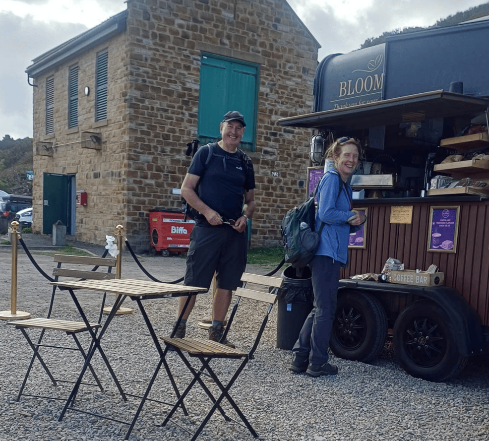 Two hikers standing at a coffee cart named 'Bloom', smiling and enjoying their visit. A stone building is in the background, along with outdoor seating.