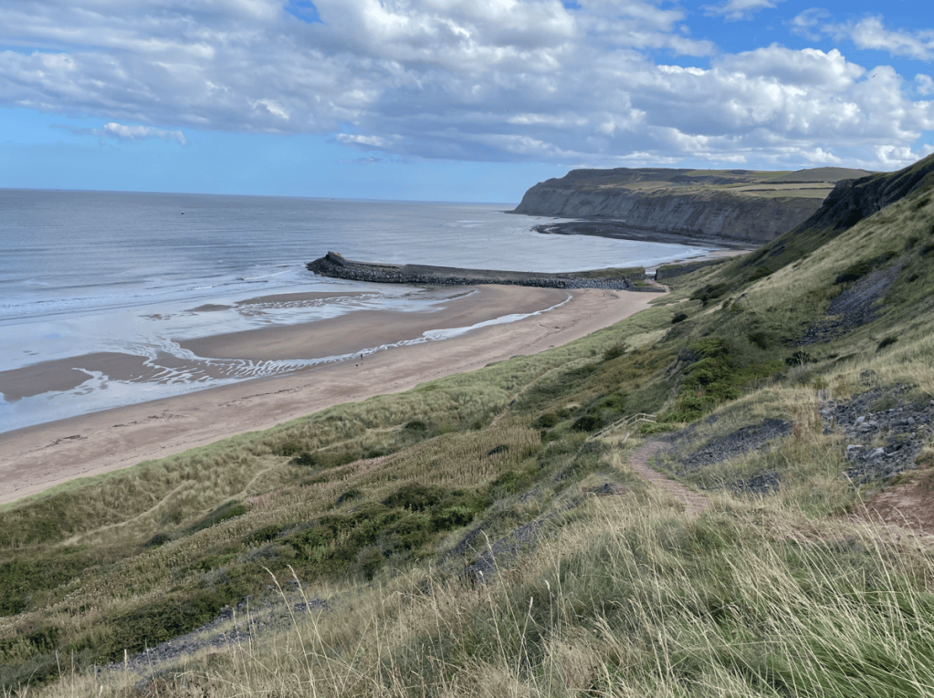A scenic view of a sandy beach bordered by cliffs and a calm sea, with a rocky jetty extending into the water under a partly cloudy sky.