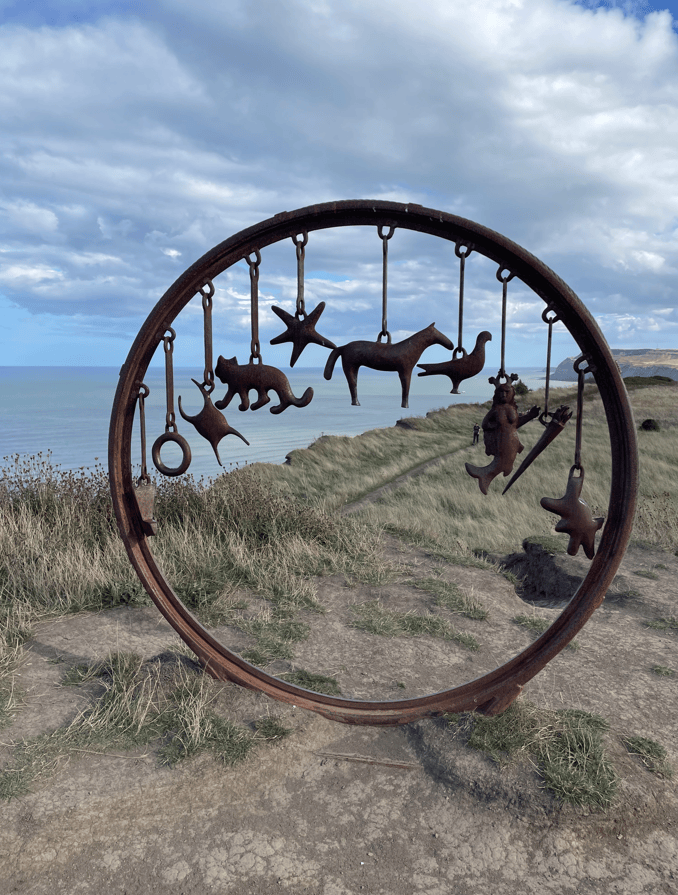 A circular metal sculpture featuring various animal and character shapes hanging from it, set against a backdrop of cliffs and the sea under a cloudy sky.