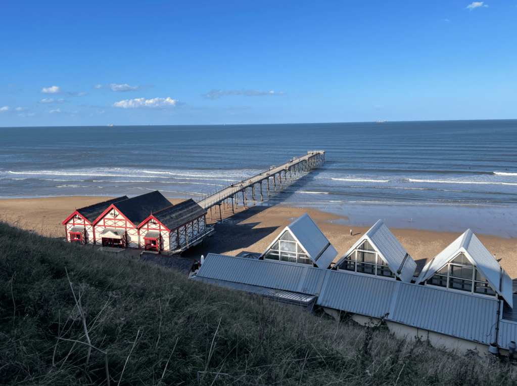 A scenic view of a beach with a wooden pier extending into the sea, alongside traditional beach huts with red accents and modern glass-roofed buildings.