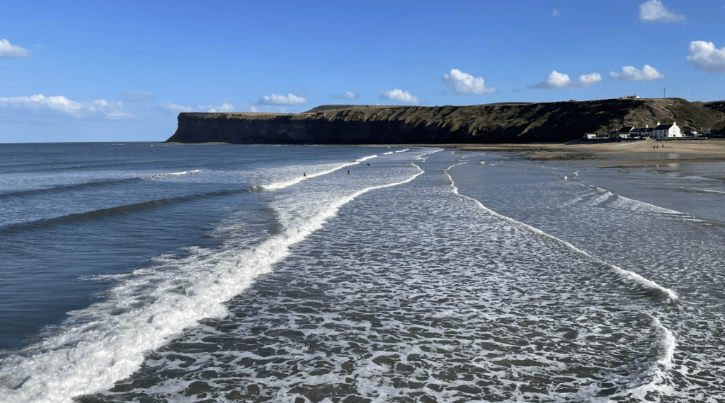 A serene beach scene featuring gentle waves lapping at the shore, framed by a cliffside and a clear blue sky with scattered clouds.