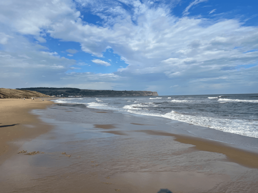 A scenic beach view with golden sand, gentle waves, and a cloudy blue sky, featuring distant cliffs and people walking along the shoreline.