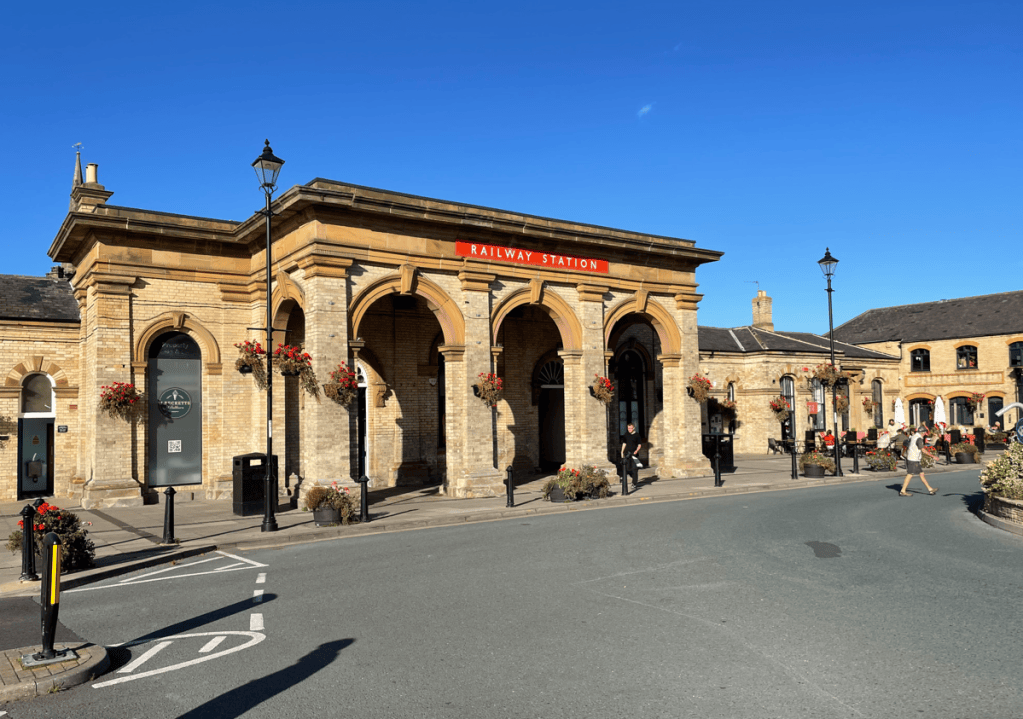 View of a historic railway station building with arched entrances and flower baskets, set against a clear blue sky. People walking and parked bicycles in the foreground.