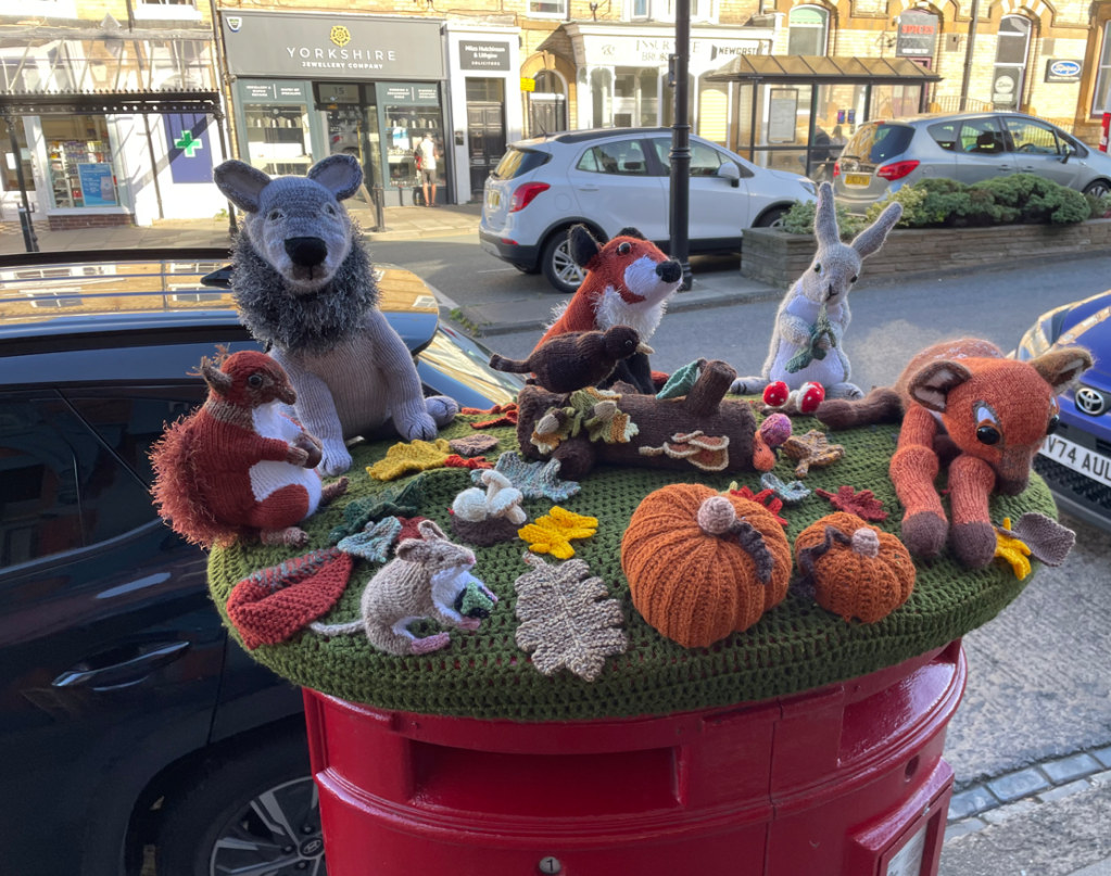 A collection of knitted animals, including a wolf, fox, rabbit, and squirrel, displayed on a green knitted surface atop a red postbox. The scene features autumn leaves, acorns, and pumpkins, creating a whimsical outdoor decoration.