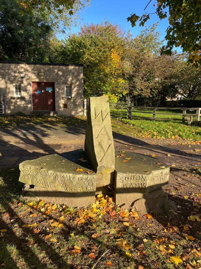 A stone monument with engravings situated in a park, featuring autumn foliage, a building in the background, and fallen leaves on the ground.