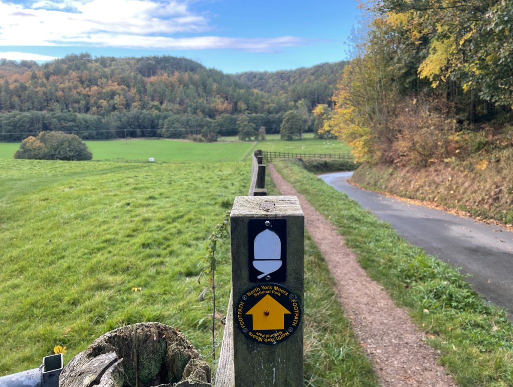 A signpost indicating a walking path towards the North York Moors, set against a backdrop of lush green fields and hills under a cloudy sky.