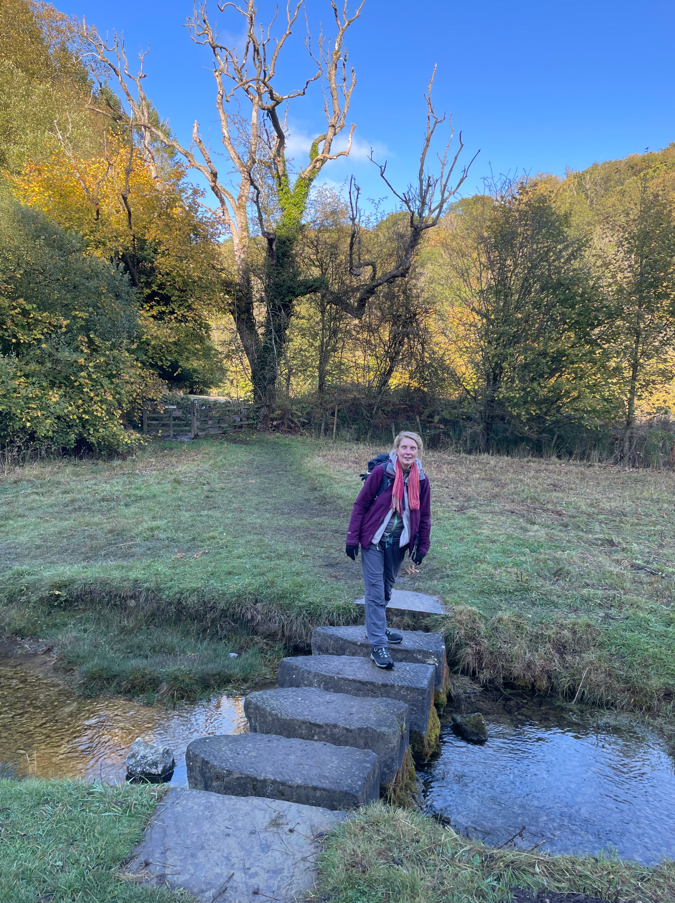 A woman standing on stone stepping stones crossing a stream, surrounded by a lush green landscape with trees and a clear blue sky.