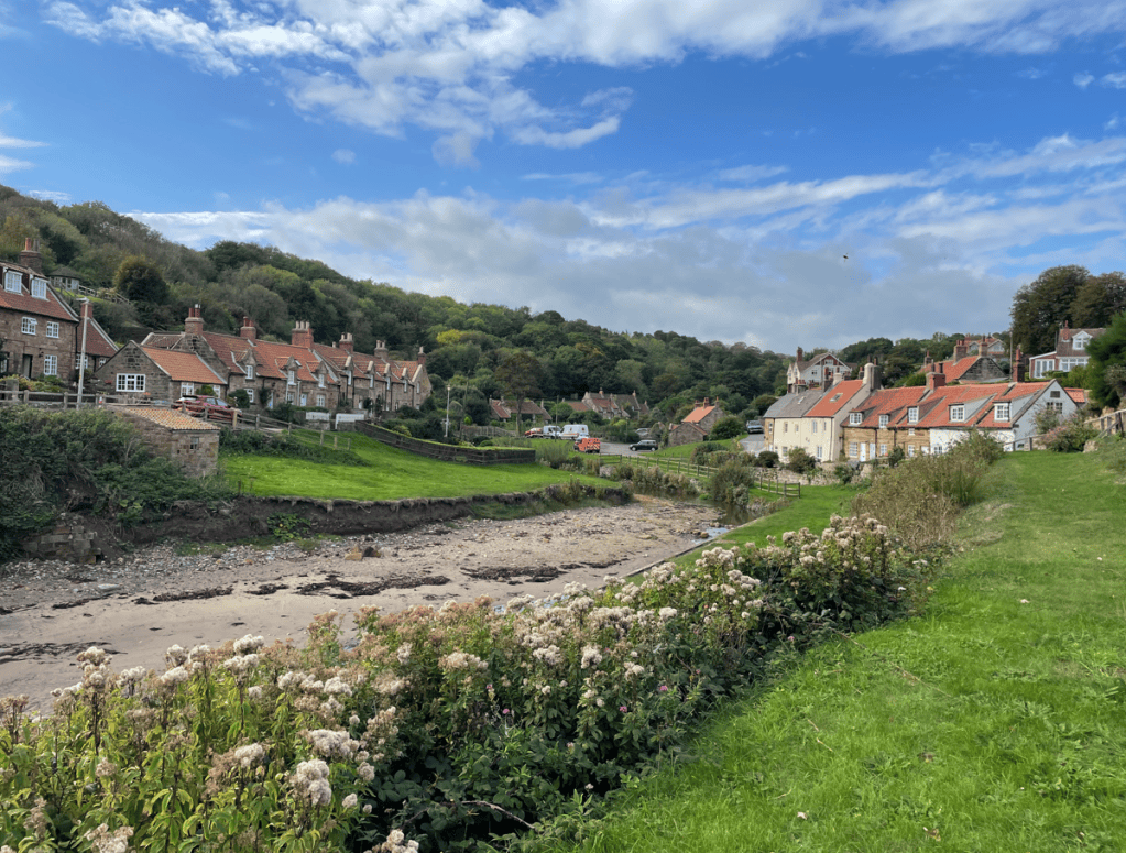 A picturesque view of a coastal village with traditional cottages, rolling green hills, and a clear blue sky.