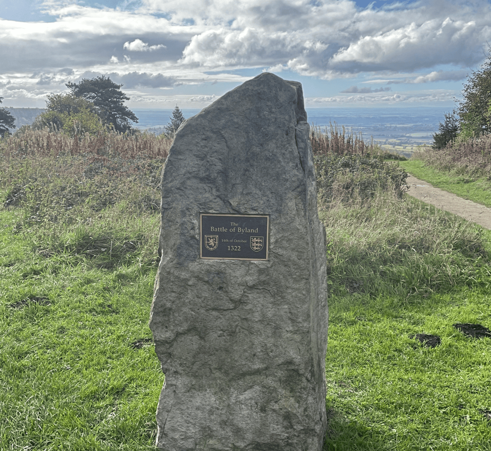 A commemorative stone marking the site of the Battle of Byland, featuring a plaque with the inscription 'The Battle of Byland 14th of October 1322', surrounded by a grassy landscape and a cloudy sky.