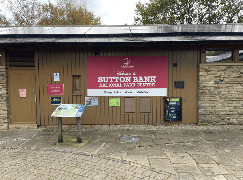Exterior view of the Sutton Bank National Park Centre with a welcoming sign, displaying information about the shop, exhibitions, and a map stand.
