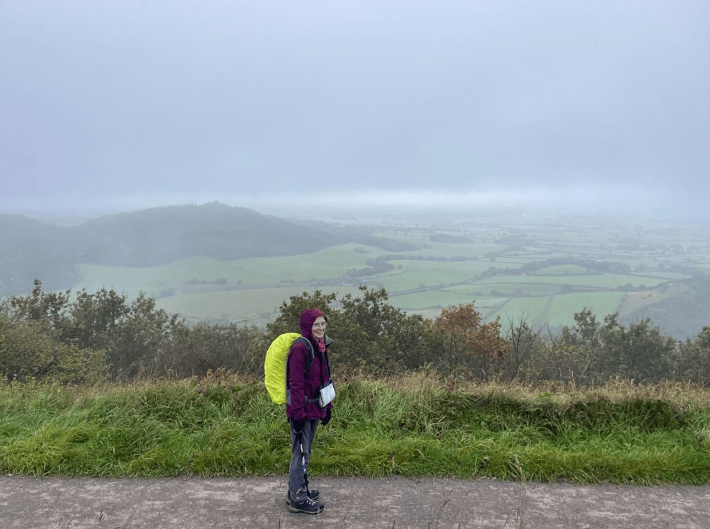 A person wearing a purple jacket and carrying a bright yellow backpack stands on a hillside overlooking a misty, green landscape.
