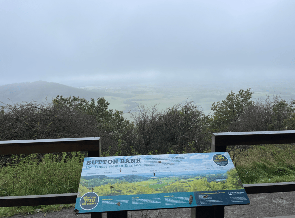 A scenic viewpoint at Sutton Bank, featuring a sign detailing local landmarks and attractions, with a misty landscape in the background.