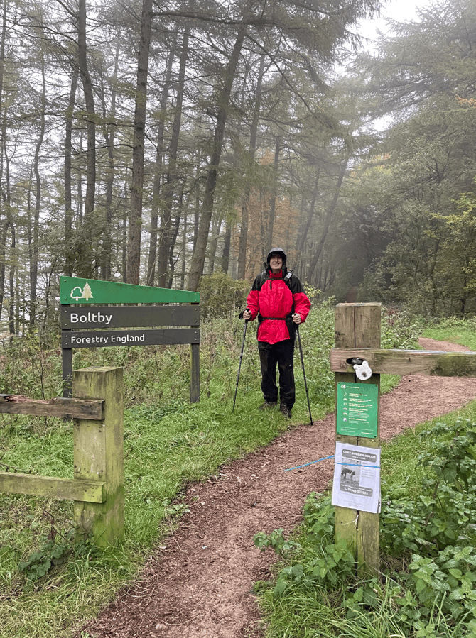 A person wearing a red rain jacket and black trousers stands at the entrance of a path marked 'Boltby, Forestry England' amidst misty trees.