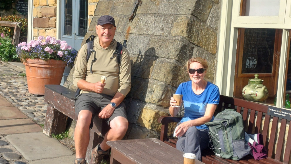 A man and woman seated on a bench outside a stone building, enjoying ice cream and coffee. The man is wearing a cap and a long-sleeve top, while the woman, in a blue t-shirt and sunglasses, smiles at the camera. Flowers in a pot are visible nearby.