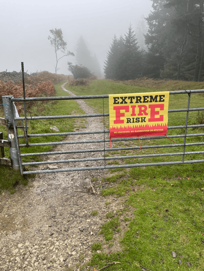 A metal gate with a bright yellow and red sign reading 'EXTREME FIRE RISK' attached to it. The sign warns against smoking, barbecues, or stoves. Behind the gate is a misty path leading into a wooded area.