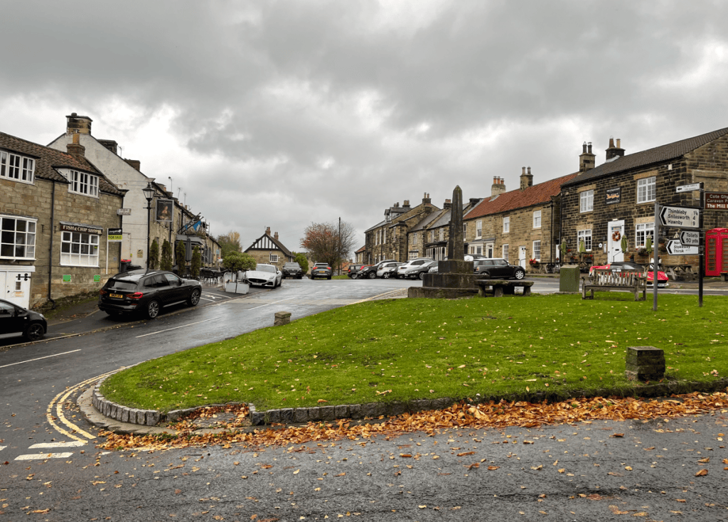 A scenic village street with traditional stone buildings and a grassy roundabout, surrounded by parked cars and autumn leaves under a cloudy sky.