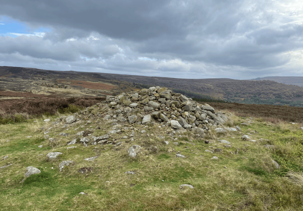 A stone cairn set against a backdrop of rolling hills and dramatic cloudy skies, with patches of moorland in the foreground.