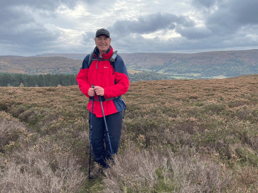 A person standing on a hillside, wearing a red jacket and holding a walking stick, with a scenic backdrop of hills and cloudy sky.