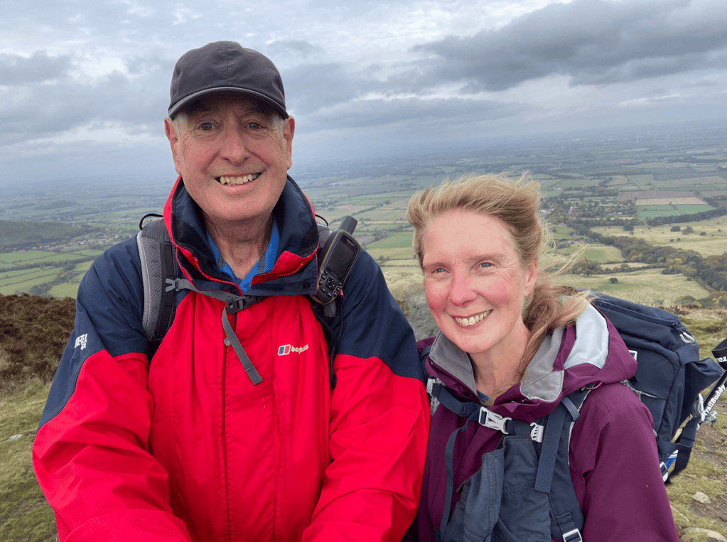 A man and a woman smiling for a selfie at the summit of a hill, with green fields and a cloudy sky in the background.