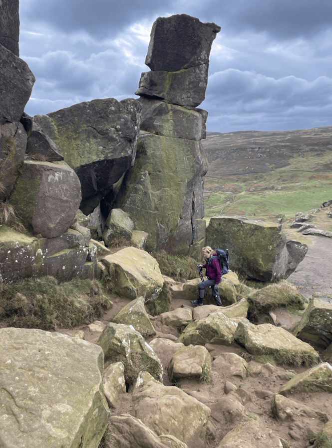 A hiker in a purple jacket resting on a rocky path surrounded by large boulders and grass, with a dramatic cloudy sky in the background.