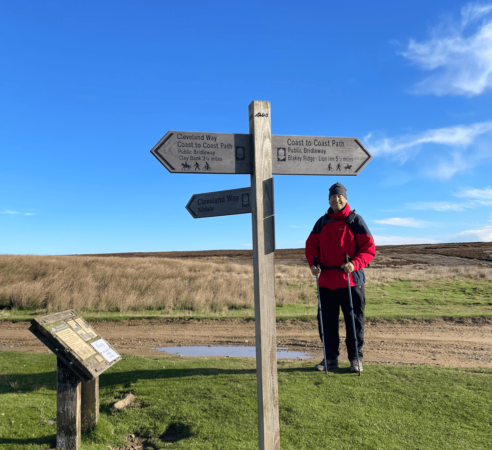 A person in a red jacket stands next to a wooden signpost with multiple directional arrows indicating walking paths, including the Cleveland Way and Coast to Coast Path, set against a blue sky and grassy background.
