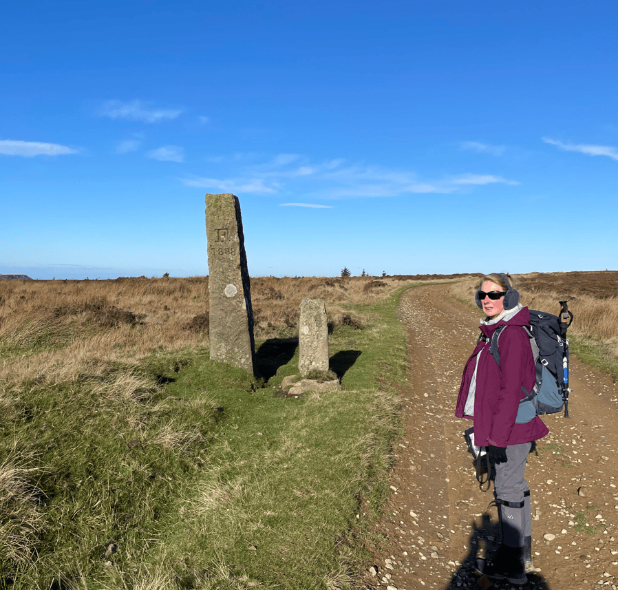 A hiker in a purple jacket stands beside two stone markers on a dirt path, with blue skies and grassland in the background.