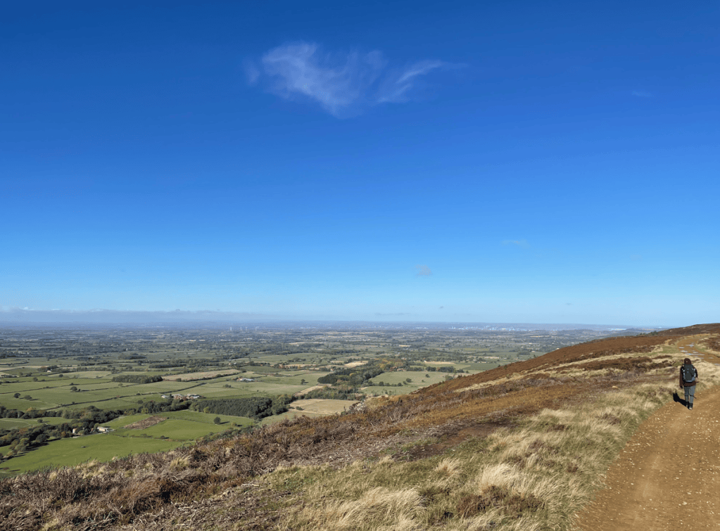 A person walking along a dirt path on a hillside, overlooking a vast green landscape and blue sky.