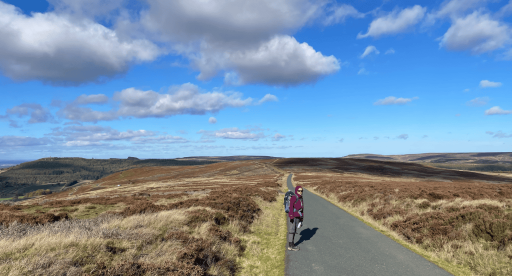 A person standing on a wide, winding road surrounded by open moorland and under a blue sky with scattered clouds.