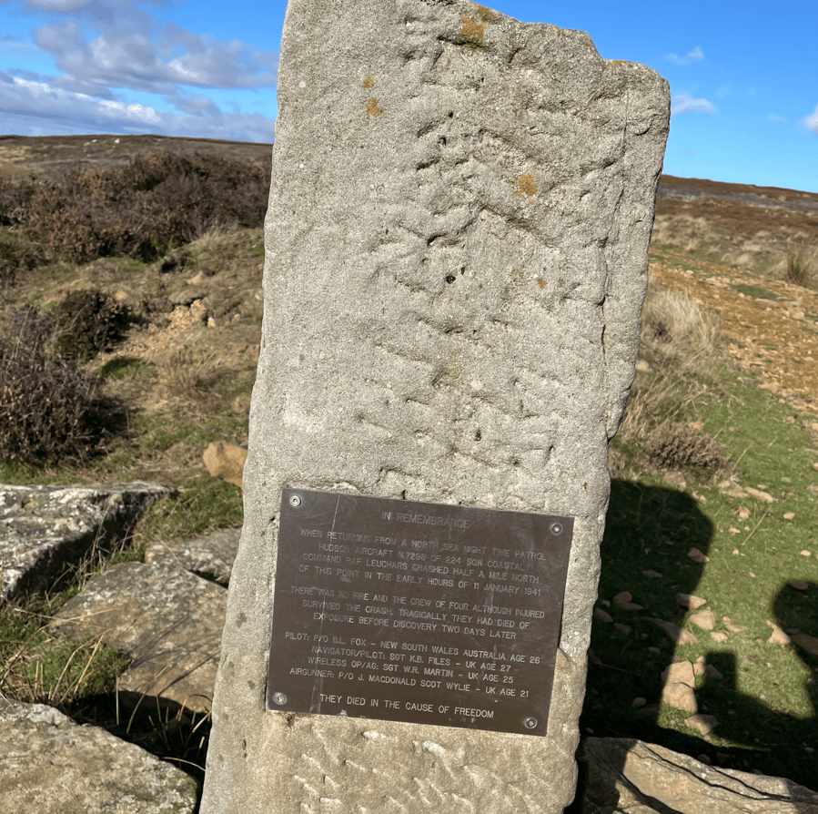 A commemorative stone memorial with an attached plaque, set against a backdrop of hills and blue skies. The plaque details the tragic crash of a Hudson aircraft in January 1941 and honours the crew members who did not survive.