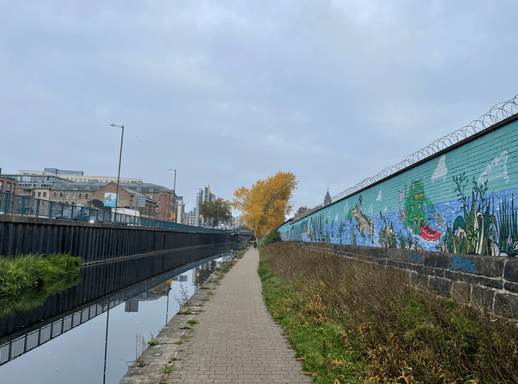 A canal walkway lined with a grassy bank, with a mural depicting aquatic life on a nearby wall, under a cloudy sky.