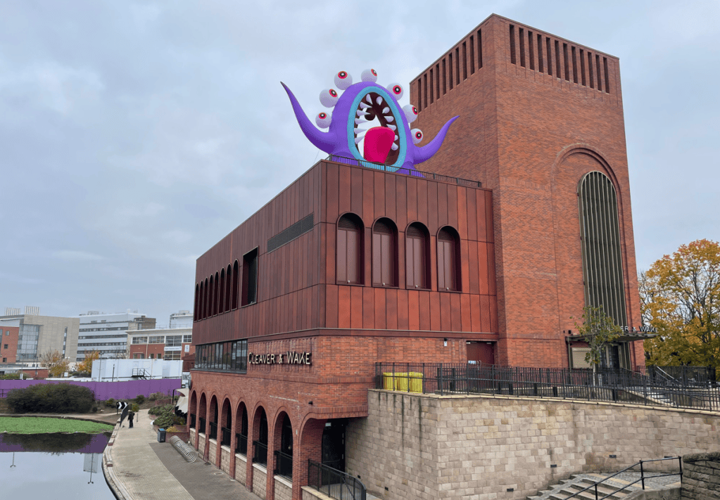 Exterior view of a modern building with a brick facade, featuring a large, colourful sculpture on the roof resembling a whimsical creature, set against a cloudy sky.